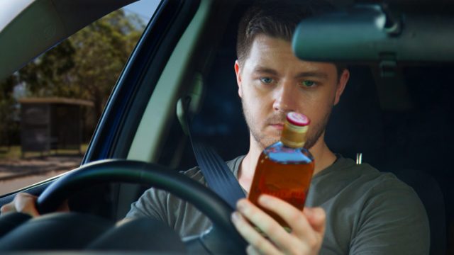 Man holds a bottle of alcohol and looks at it with one hand on the steering wheel