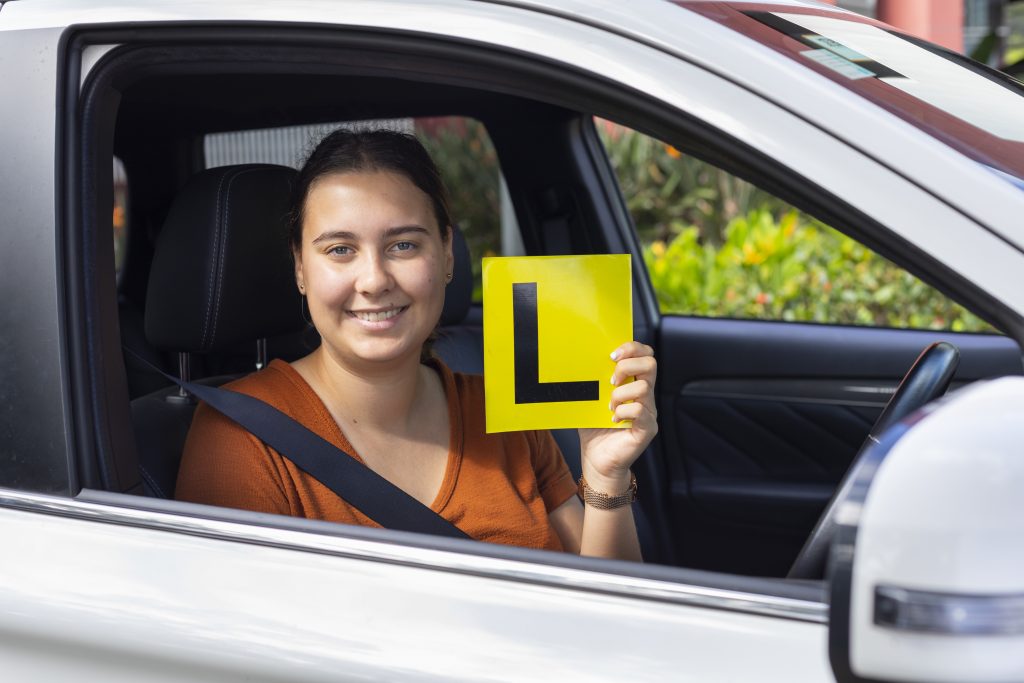 Young woman sitting in driver's seat of car holds Learner's plate up