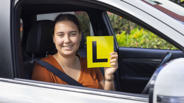 Young woman sitting in driver's seat of car holds Learner's plate up
