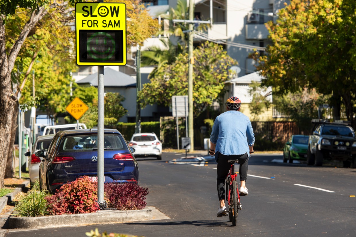 'Slow down for Sam' sign on road with a bicycle rider next to it