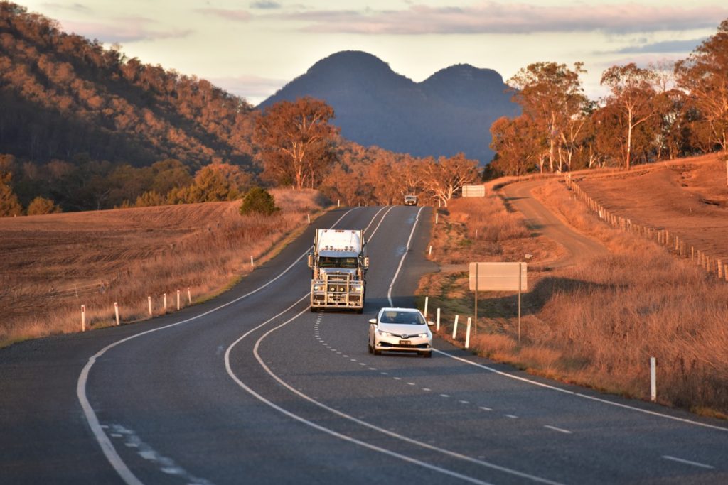 Truck follows car down long stretch of regional road surrounded by trees and mountains.