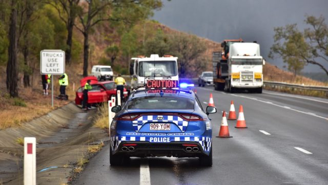 Blue road police car with flashing lights pulled over on side of wet mountain road to a crash scene involving a red car. Police and paramedic personnel surround.