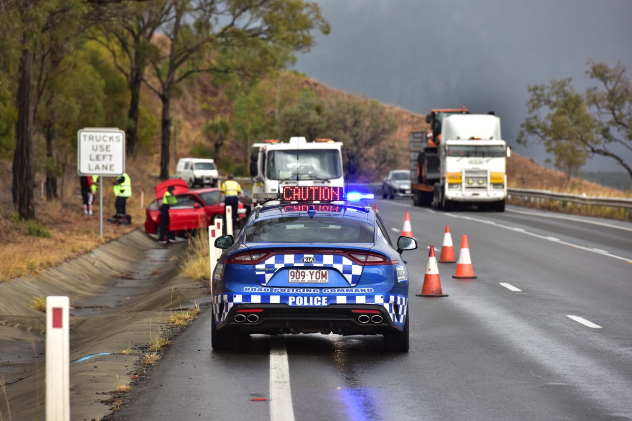 Blue road police car with flashing lights pulled over on side of wet mountain road to a crash scene involving a red car. Police and paramedic personnel surround.